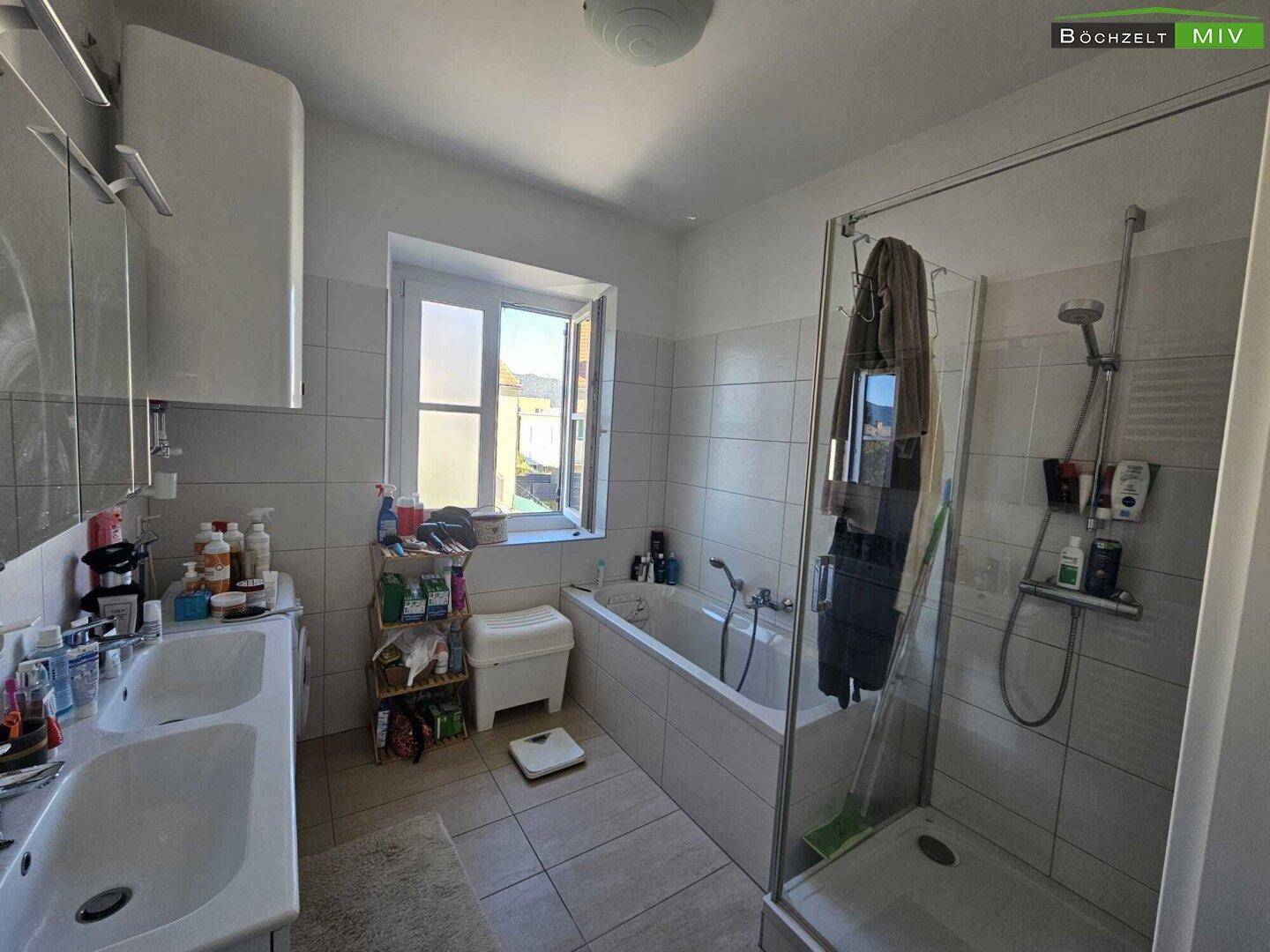 Helles Badezimmer mit Badewanne, Duschkabine, Doppelwaschbecken und Fenster.