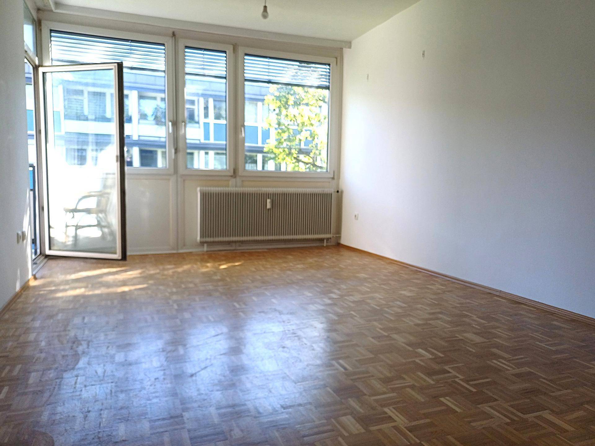 Heller Raum mit Fischgrätparkettboden, großen Fenstern und Zugang zu einem Balkon.
