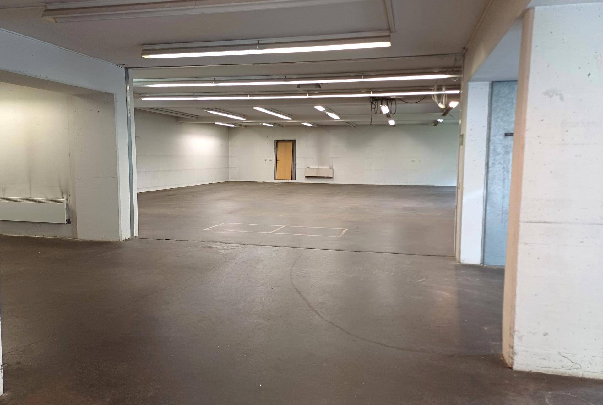 Weiter Blick in einen großen, leeren Raum mit Betonboden und einer einzelnen Tür am Ende.