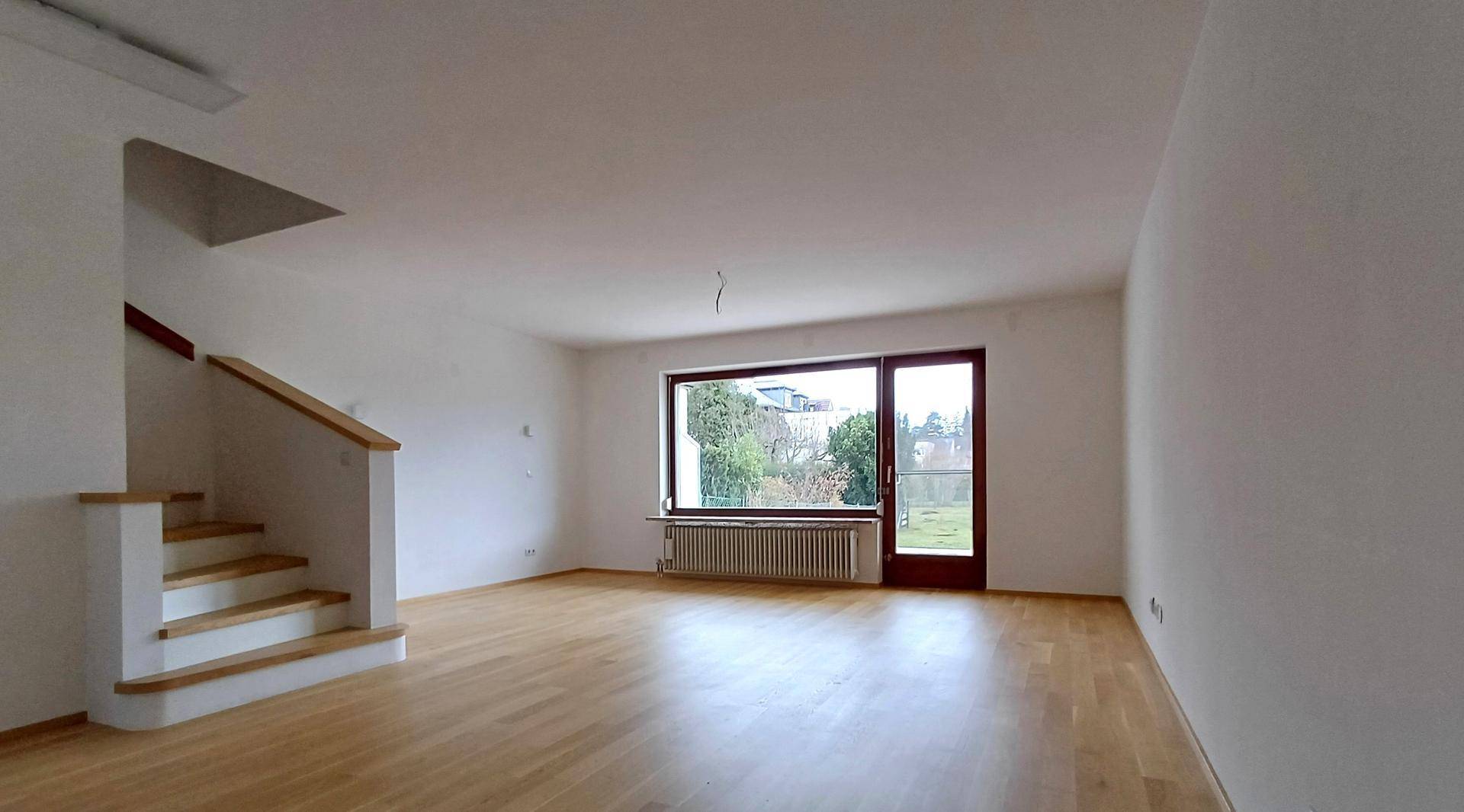 Geräumiges Wohnzimmer mit hellem Parkettboden, großem Fenster und Treppenzugang.