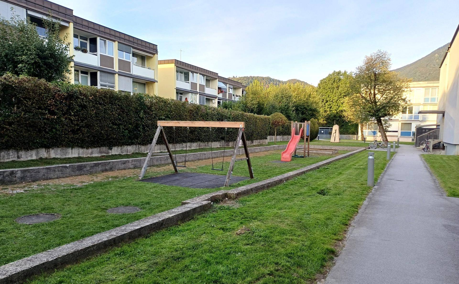 Kinderspielplatz mit Schaukel und Rutsche auf einer Grünfläche, umgeben von Wohngebäuden.