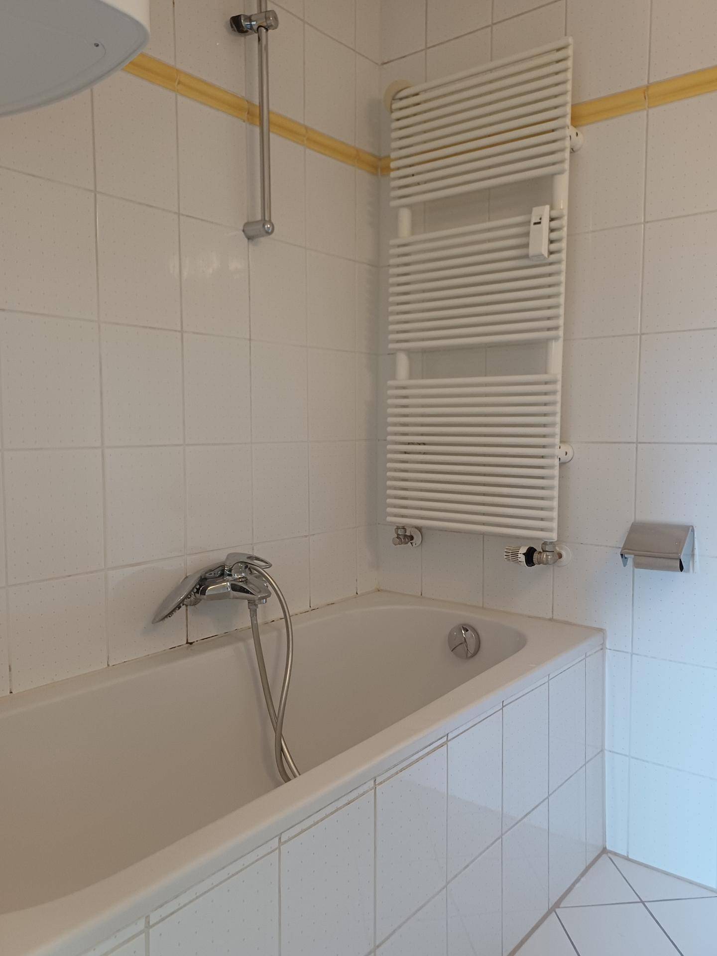 Badezimmer mit Badewanne und beheiztem Handtuchhalter, umgeben von weißen Fliesen.