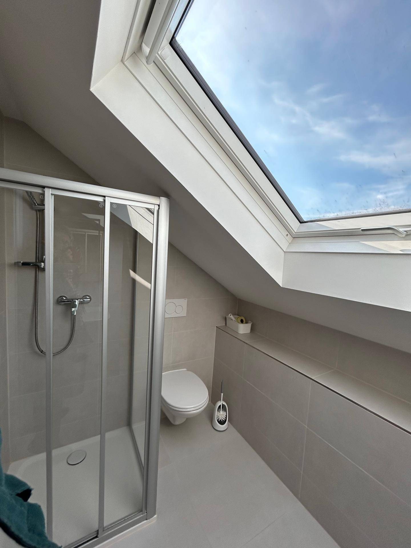 Modernes Badezimmer im Dachgeschoss mit Dusche, WC und großem Dachfenster.