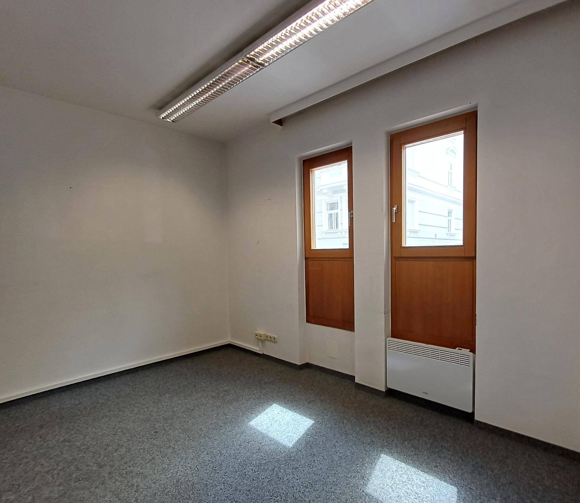 Kleiner Büroraum mit Teppichboden, zwei Fenstern und Deckenbeleuchtung.