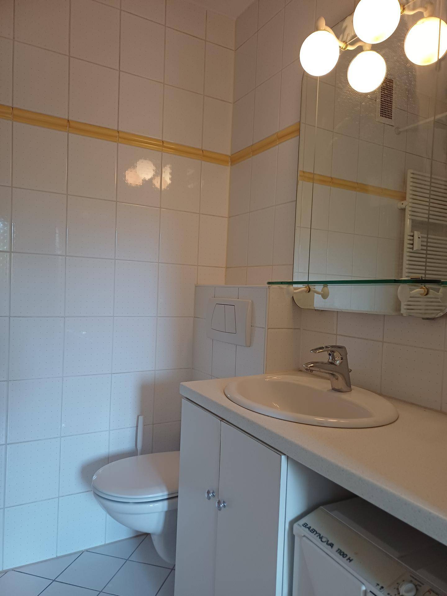 Badezimmer mit WC, Waschbecken und Spiegel, ausgestattet mit weißen Fliesen und gelber Bordüre.