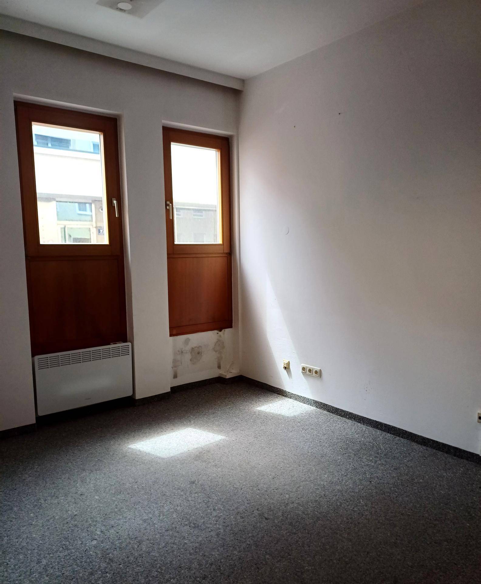 Leerer Büroraum mit Teppichboden, zwei Fenstern und sichtbaren Gebrauchsspuren an der Wand.