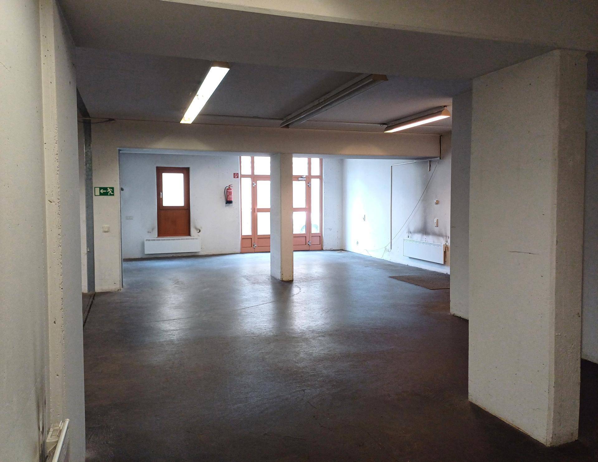 Großer, leerer Raum mit Betonboden, tragenden Säulen und Fenstern im Hintergrund.