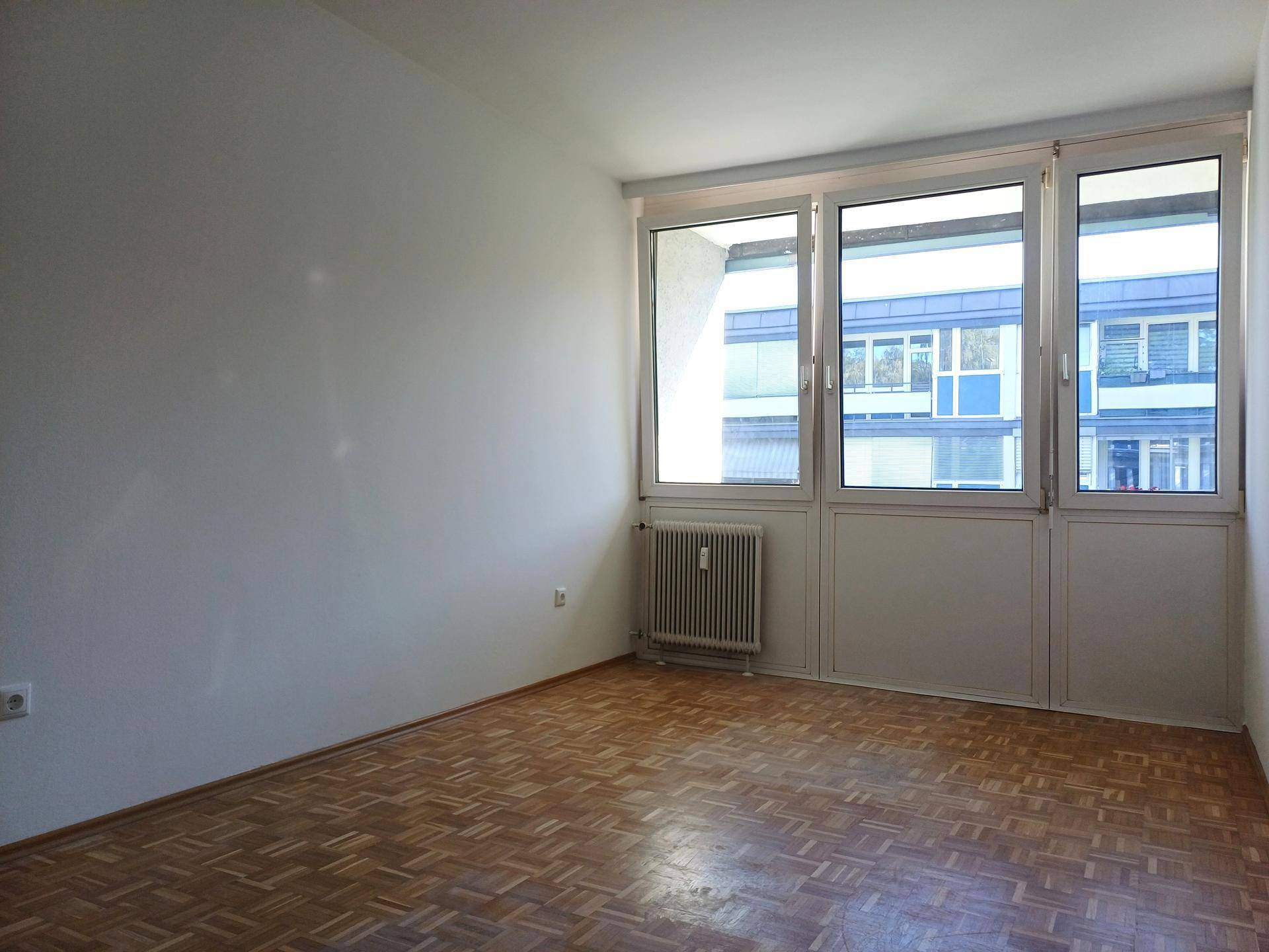 Leeres Schlafzimmer mit Parkettboden und großen Fenstern, die viel Tageslicht hereinlassen.