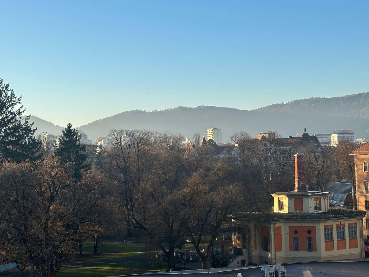 Weitläufiger Blick über einen Stadtpark mit kahlen Bäumen und Bergen im Hintergrund unter klarem Himmel.