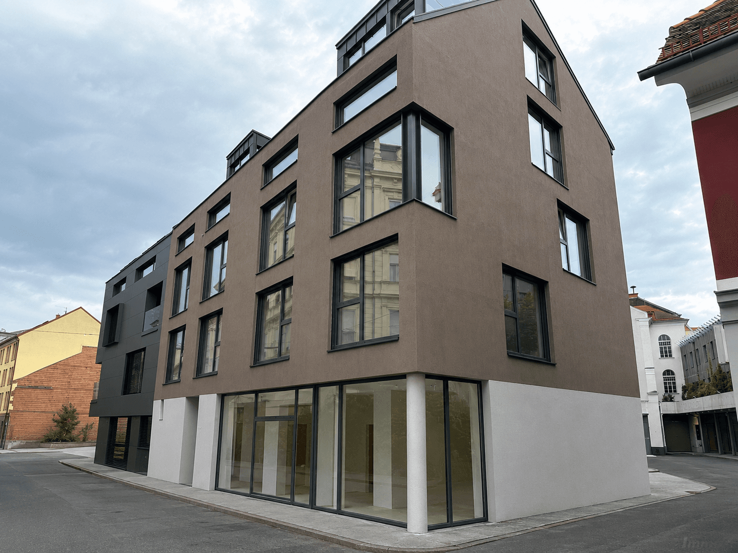 Modernes Neubaugebäude mit brauner Fassade, großen Fenstern und Geschäftsflächen im Erdgeschoss.
