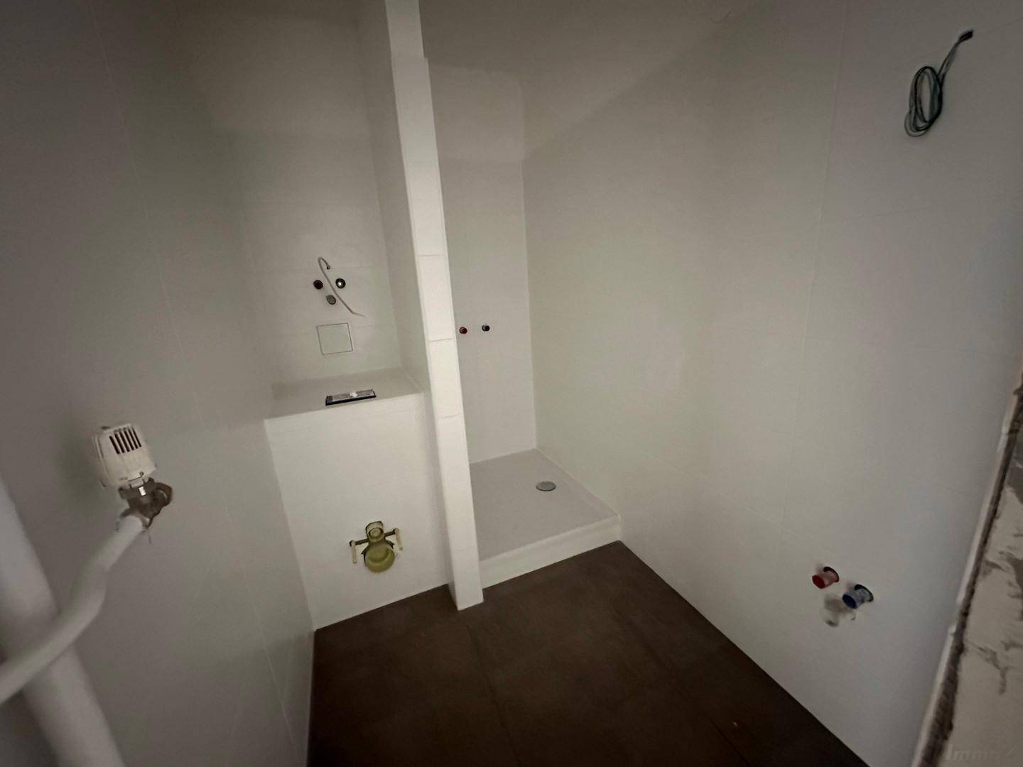 Unfertiges Badezimmer mit vorbereiteten Anschlüssen für eine Dusche und dunklem Fliesenboden.