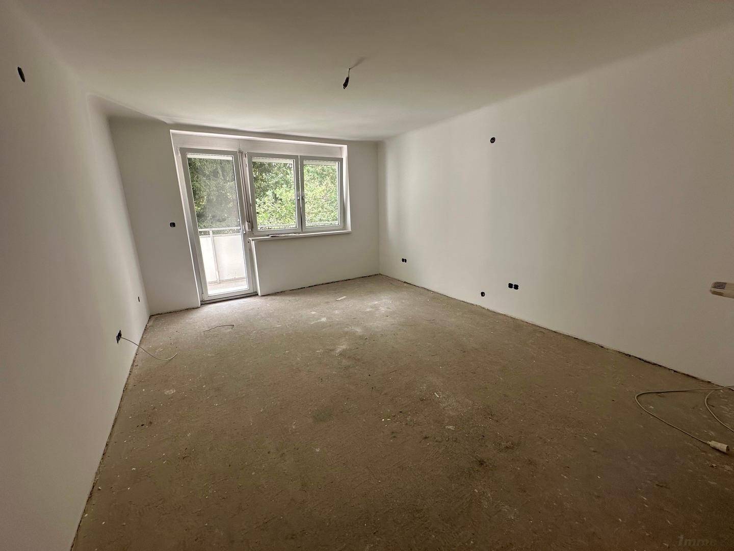 Leerer Raum mit weißen Wänden, unfertigem Boden und Fenster mit Blick ins Grüne.