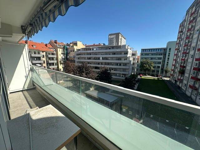 Heller Balkon mit Glasgeländer und Blick auf umliegende Gebäude und eine Grünfläche.
