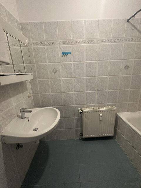 Badezimmer mit hellgrauen Wandfliesen, blauen Bodenfliesen, Waschbecken und Badewanne.