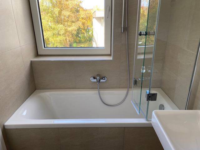 Helles Badezimmer mit Badewanne, Duschwand und Fenster mit Blick ins Grüne.