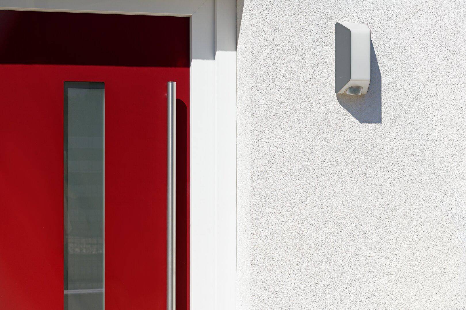 Rote Eingangstür mit langem Griff und moderner Außenbeleuchtung an der weißen Fassade.