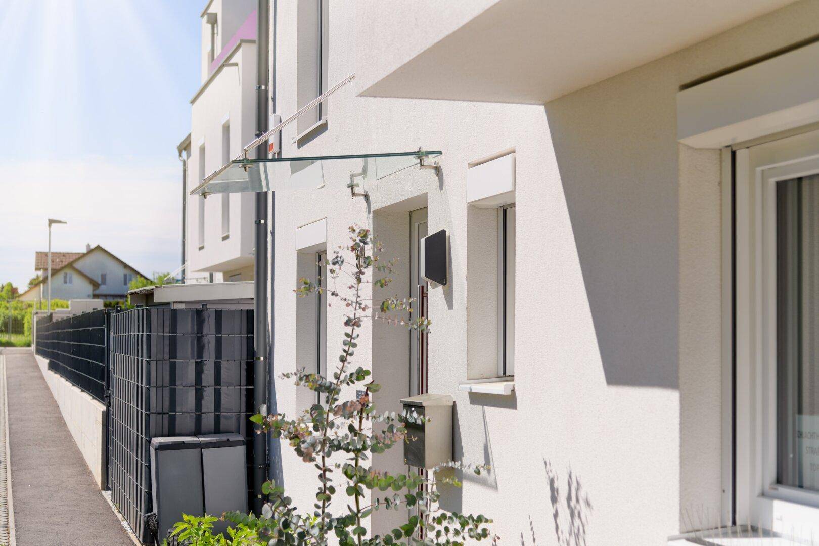 Moderner Hauseingang mit Glasvordach und Briefkasten an der weißen Fassade.