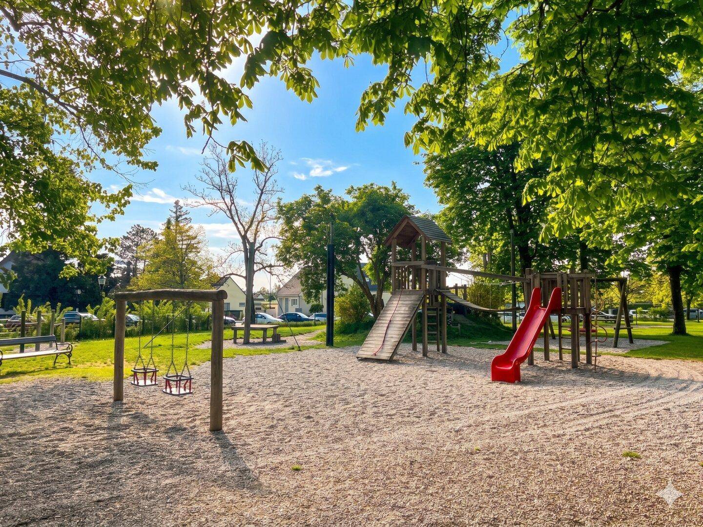 Sonniger Spielplatz mit Schaukeln, Rutsche und Sandbereich unter Bäumen.