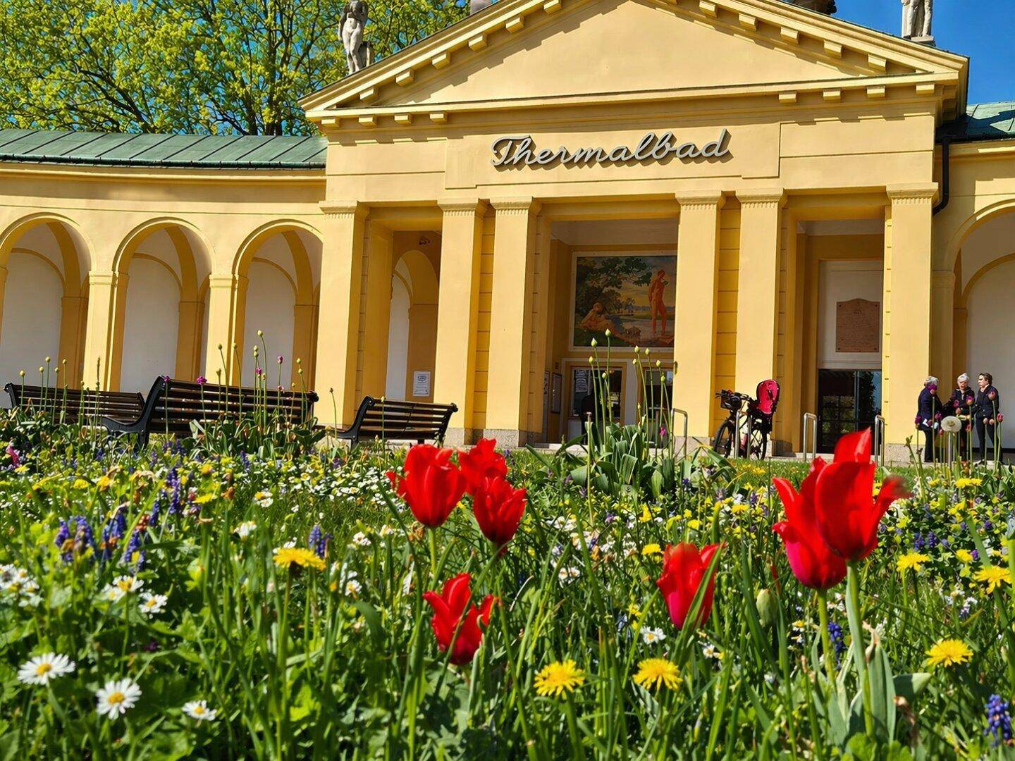 Historisches Thermalbad-Gebäude mit blühenden Blumenbeeten im Vordergrund.