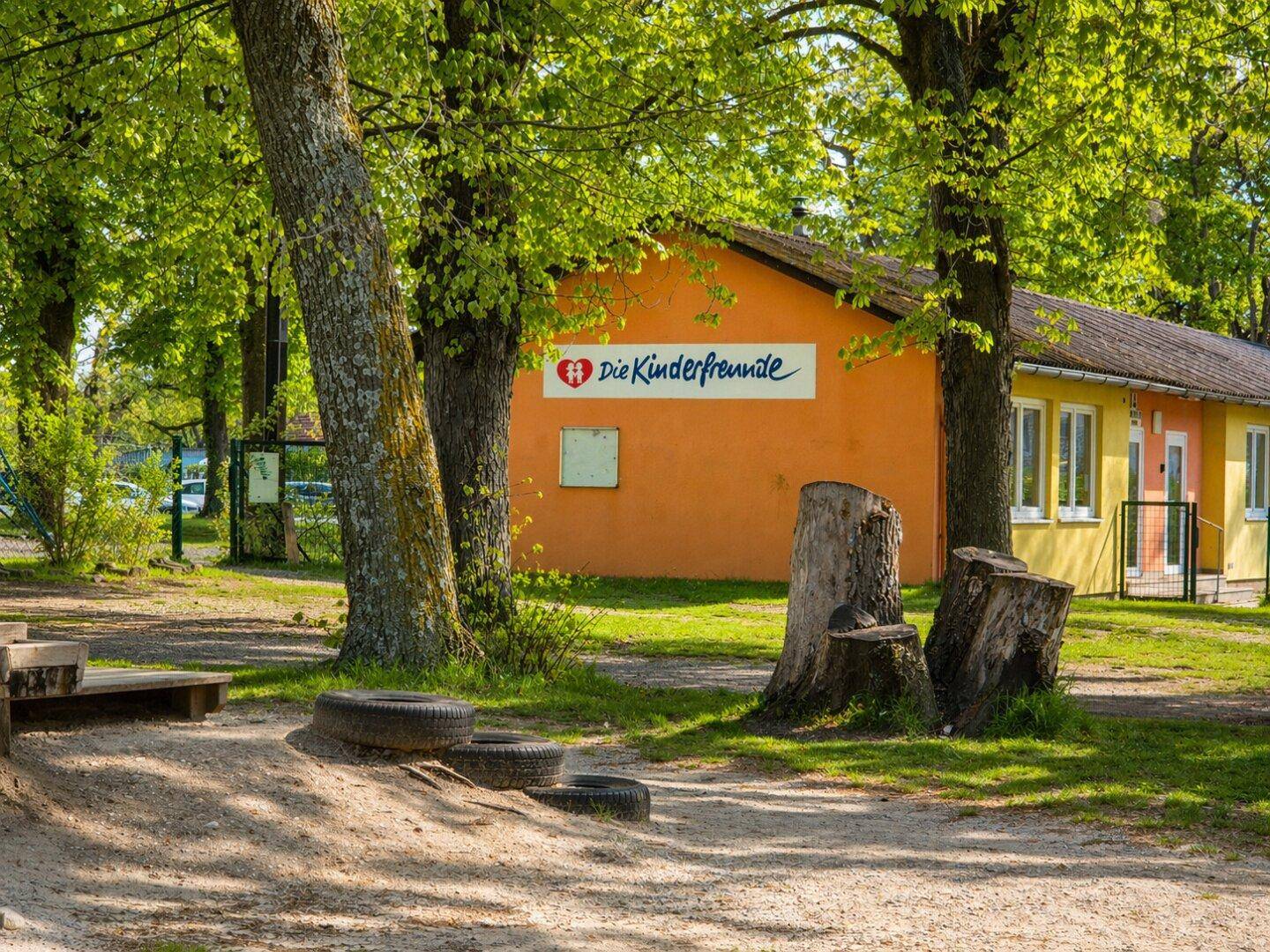 Buntes Gebäude "Die Kinderfreunde" umgeben von Bäumen und einem sandigen Bereich mit Baumstümpfen.