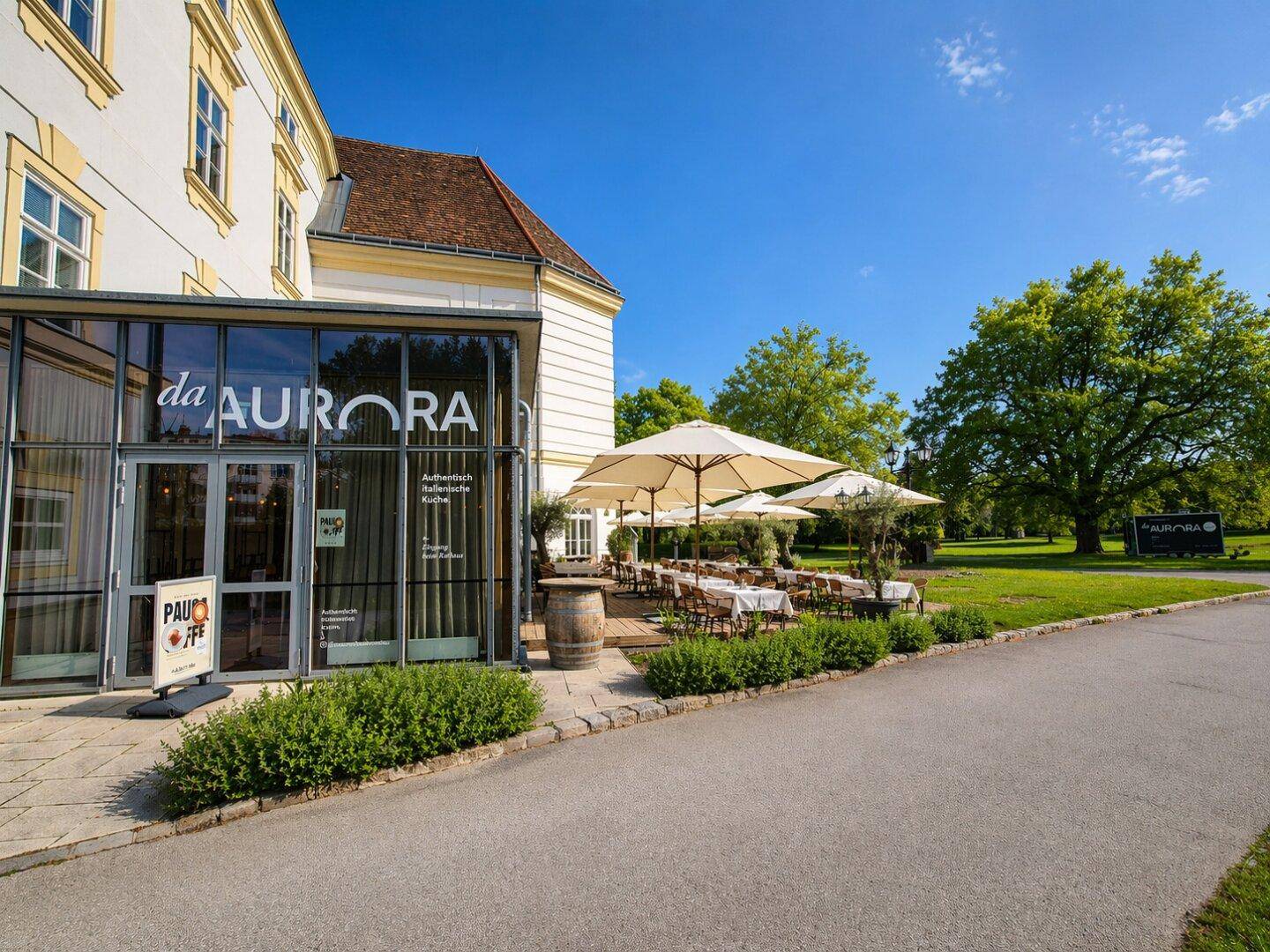 Restaurant "da AURORA" mit Glasfassade und gemütlichem Außenbereich unter großen Sonnenschirmen.