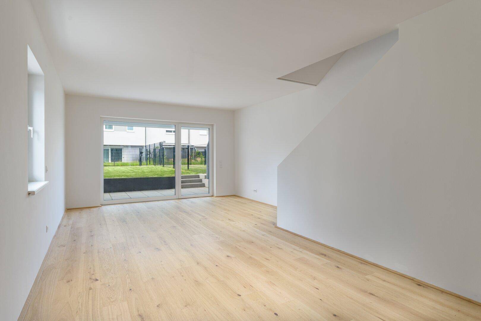 Heller, leerer Wohnraum mit hellem Holzfußboden und Zugang zu Terrasse und Garten.