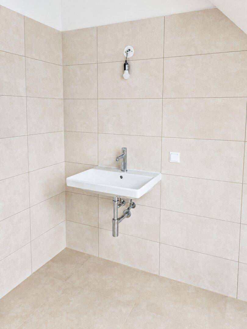 Funktionales Badezimmer mit wandmontiertem Waschbecken und hellen, beigefarbenen Fliesen an Wänden und Boden.