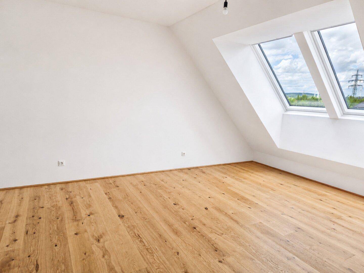 Lichtdurchflutetes Zimmer im Dachgeschoss mit Holzboden und zwei großen Dachfenstern.
