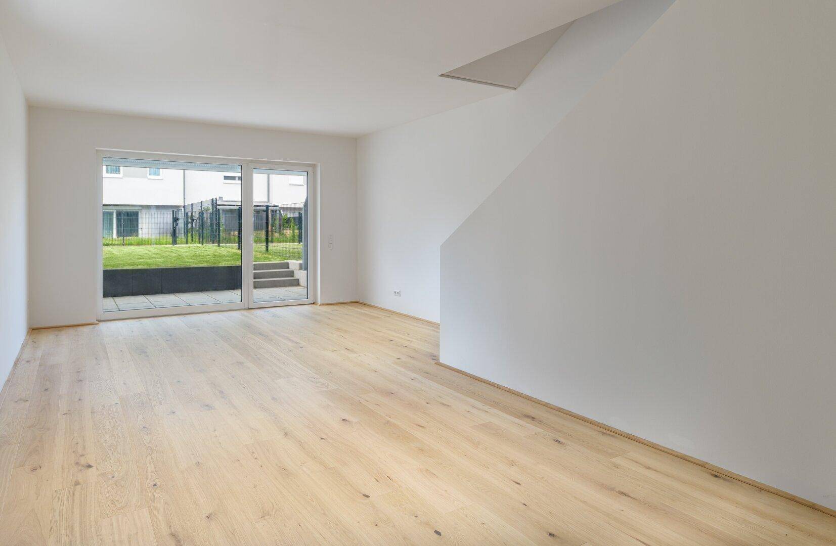 Heller, leerer Wohnraum mit Holzboden und Zugang zu einem kleinen Gartenbereich.