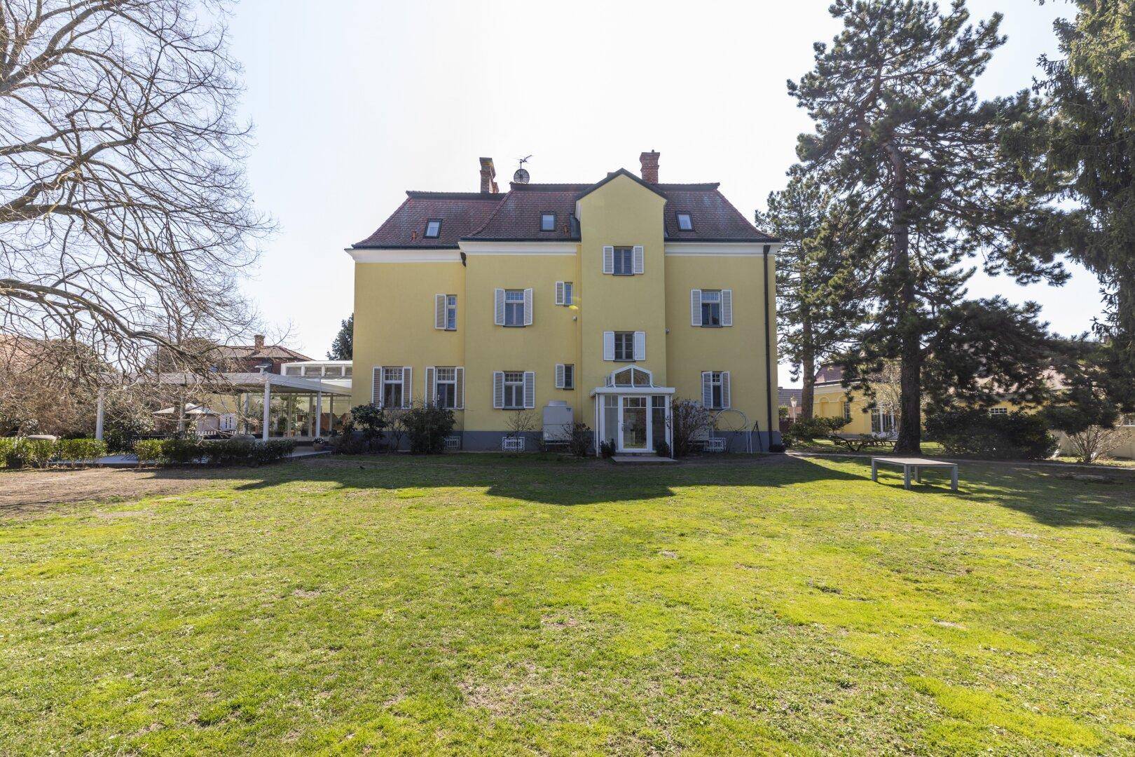 Rückansicht der gelben Villa mit weitläufigem Rasen und hohen Bäumen im Garten.