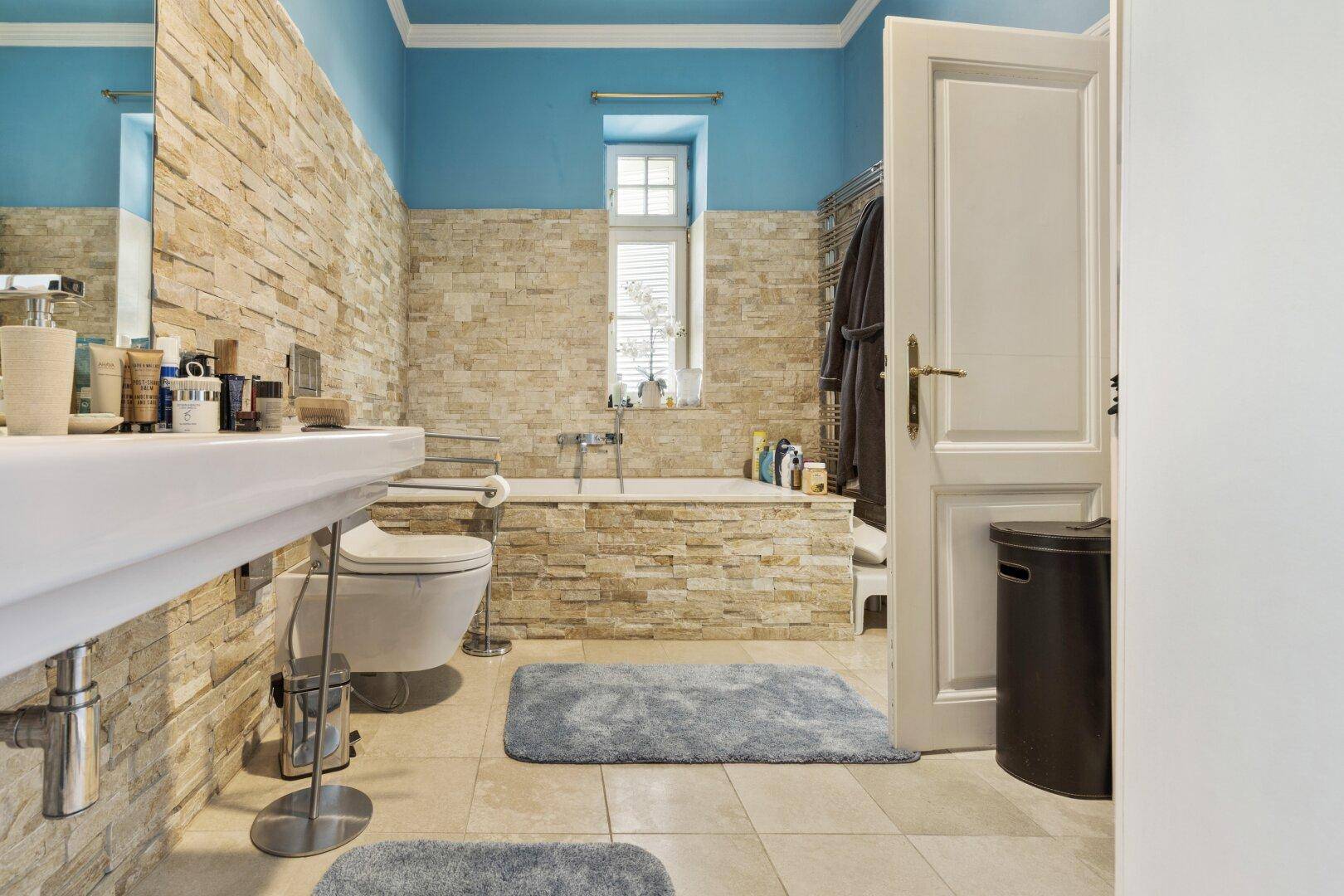 Modernes Badezimmer mit blauen Wänden, Steinverkleidung, Badewanne und Fenster.