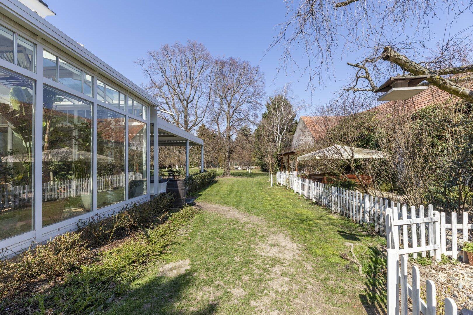 Weitläufiger Gartenbereich mit Gewächshaus, weißem Zaun und hohen Bäumen.