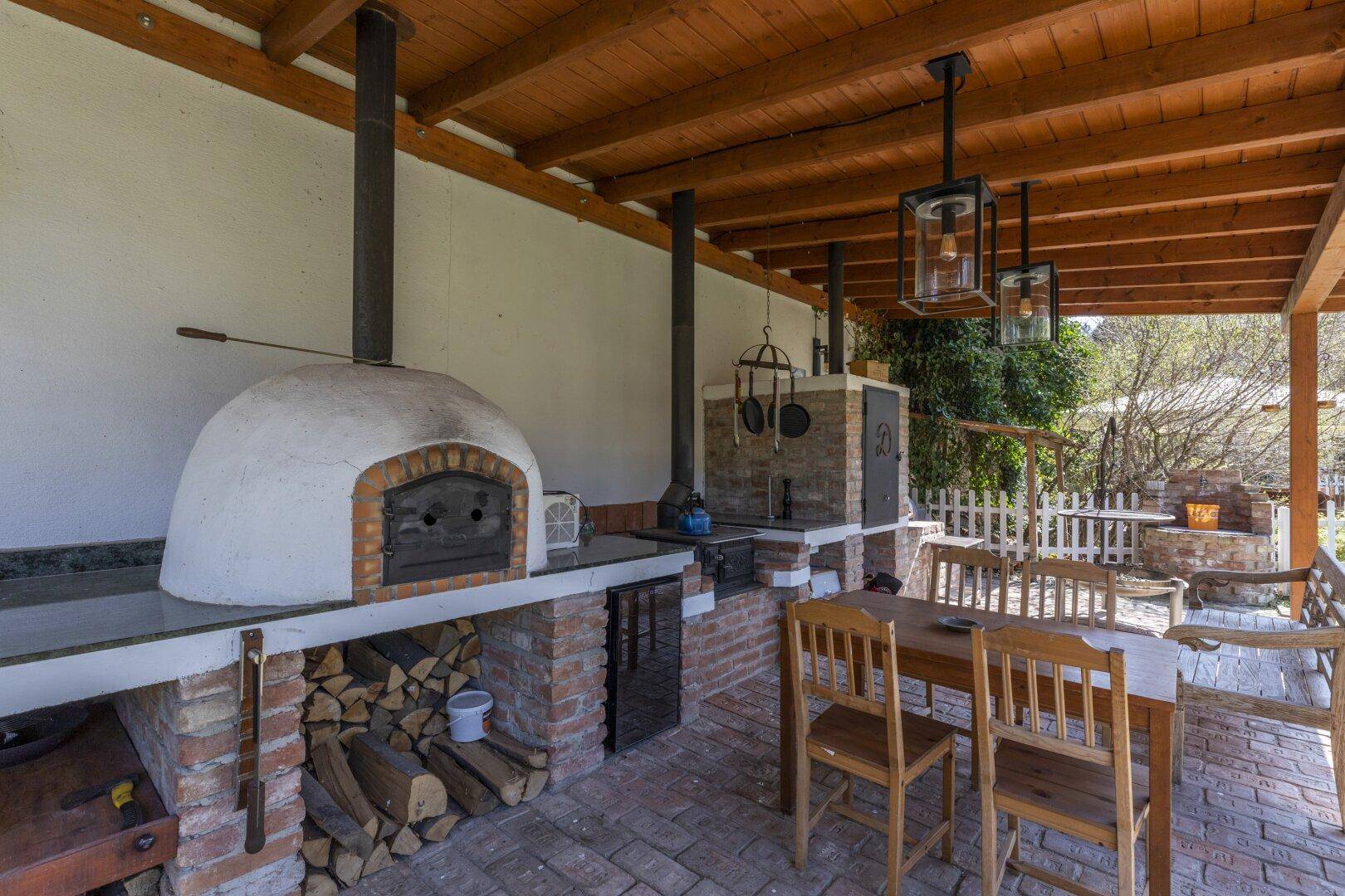 Überdachter Außenbereich mit Pizzaofen, Grillplatz und Esstisch aus Holz.