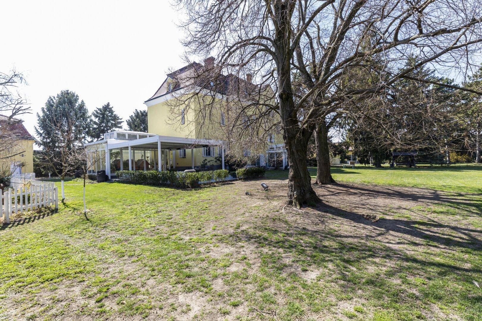 Gelbe Villa mit großem Garten und einem Wintergarten, umgeben von alten Bäumen und Rasenflächen.