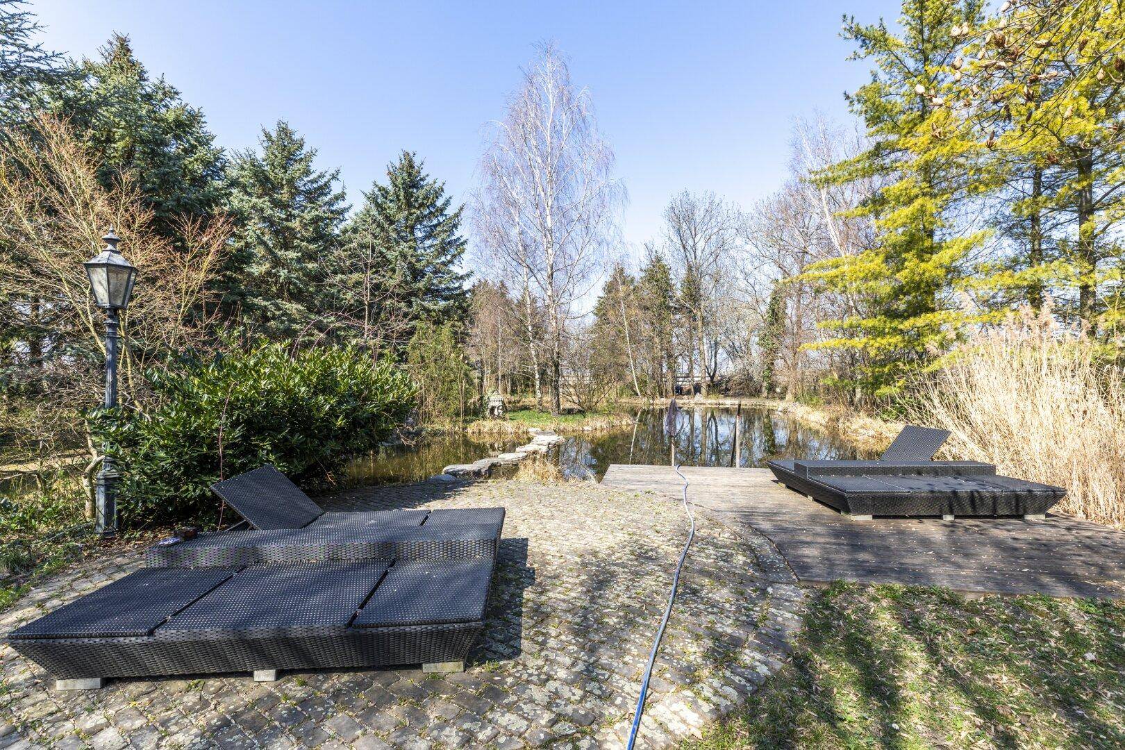 Weitläufiger Gartenbereich mit Teich, Liegeflächen und üppiger Vegetation unter blauem Himmel.