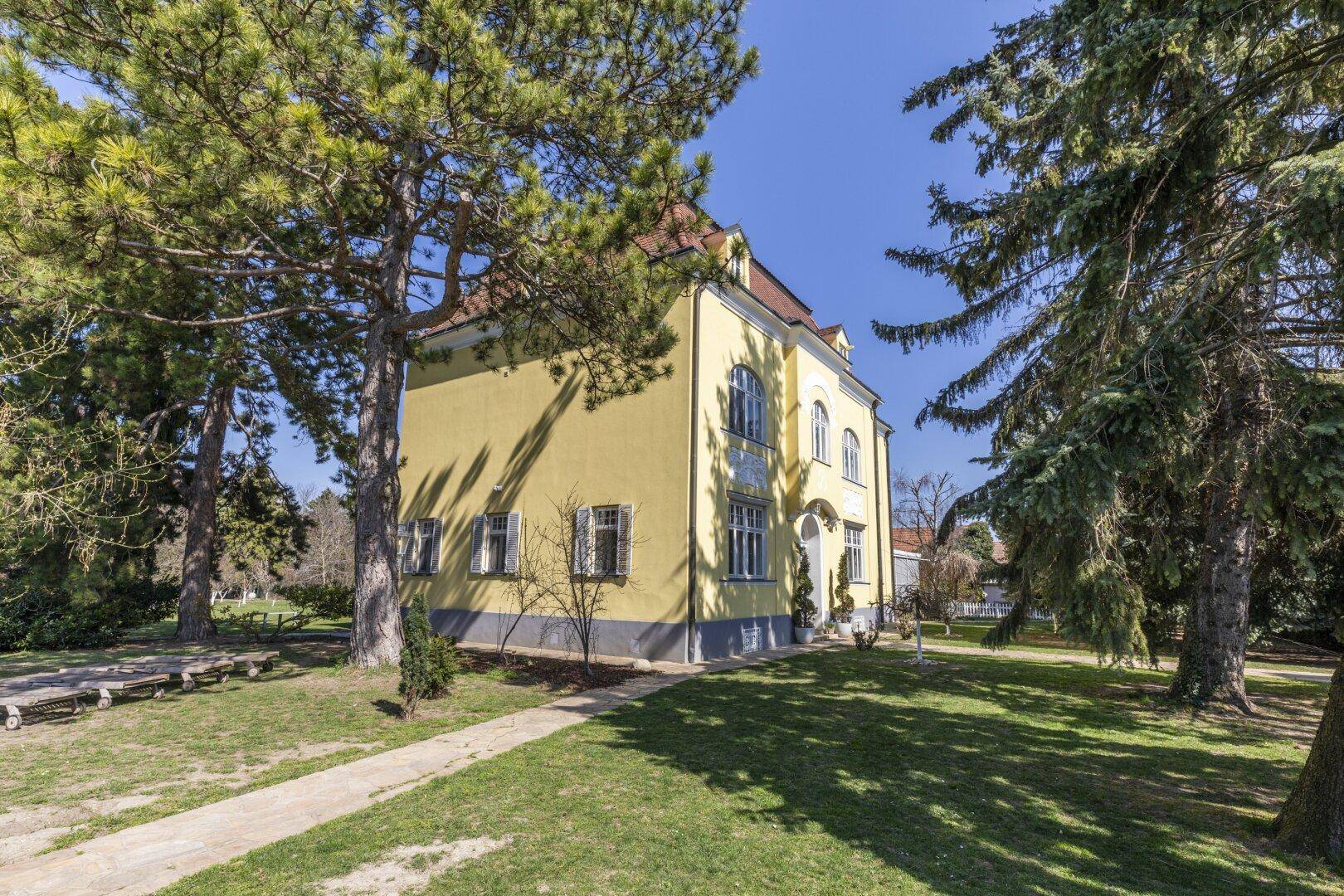 Prächtige gelbe Villa mit gepflegtem Vorgarten und hohen Bäumen unter blauem Himmel.