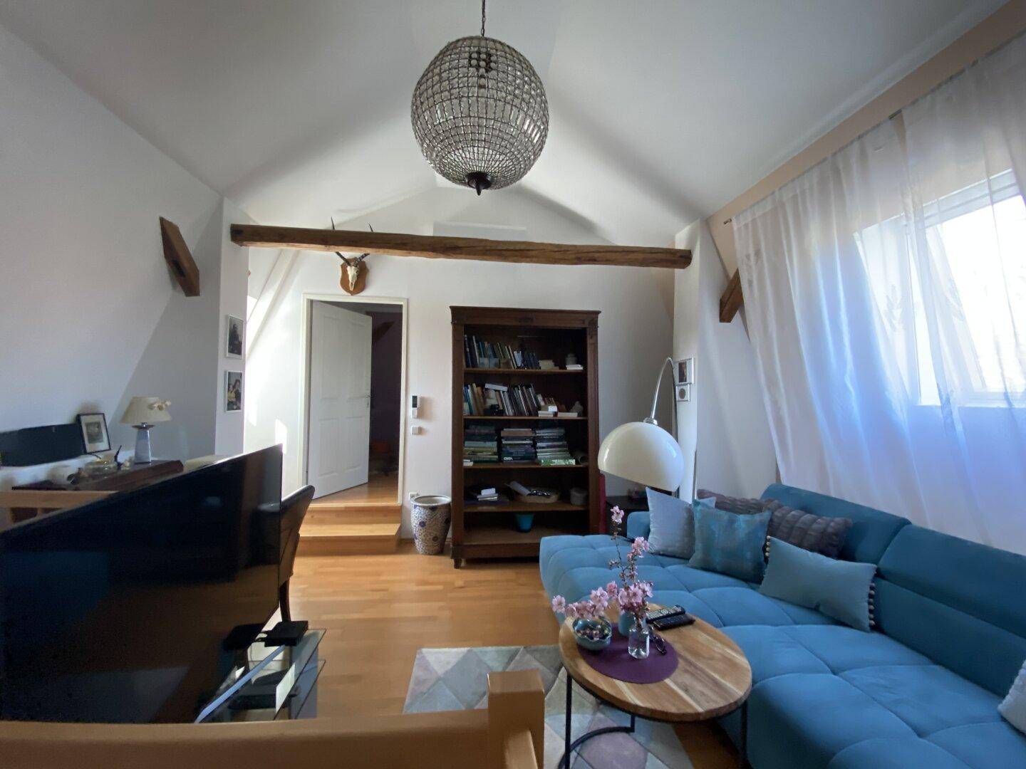 Helles Wohnzimmer mit Dachschrägen, Holzbalken und einem gemütlichen Sofa unter einem Kronleuchter.