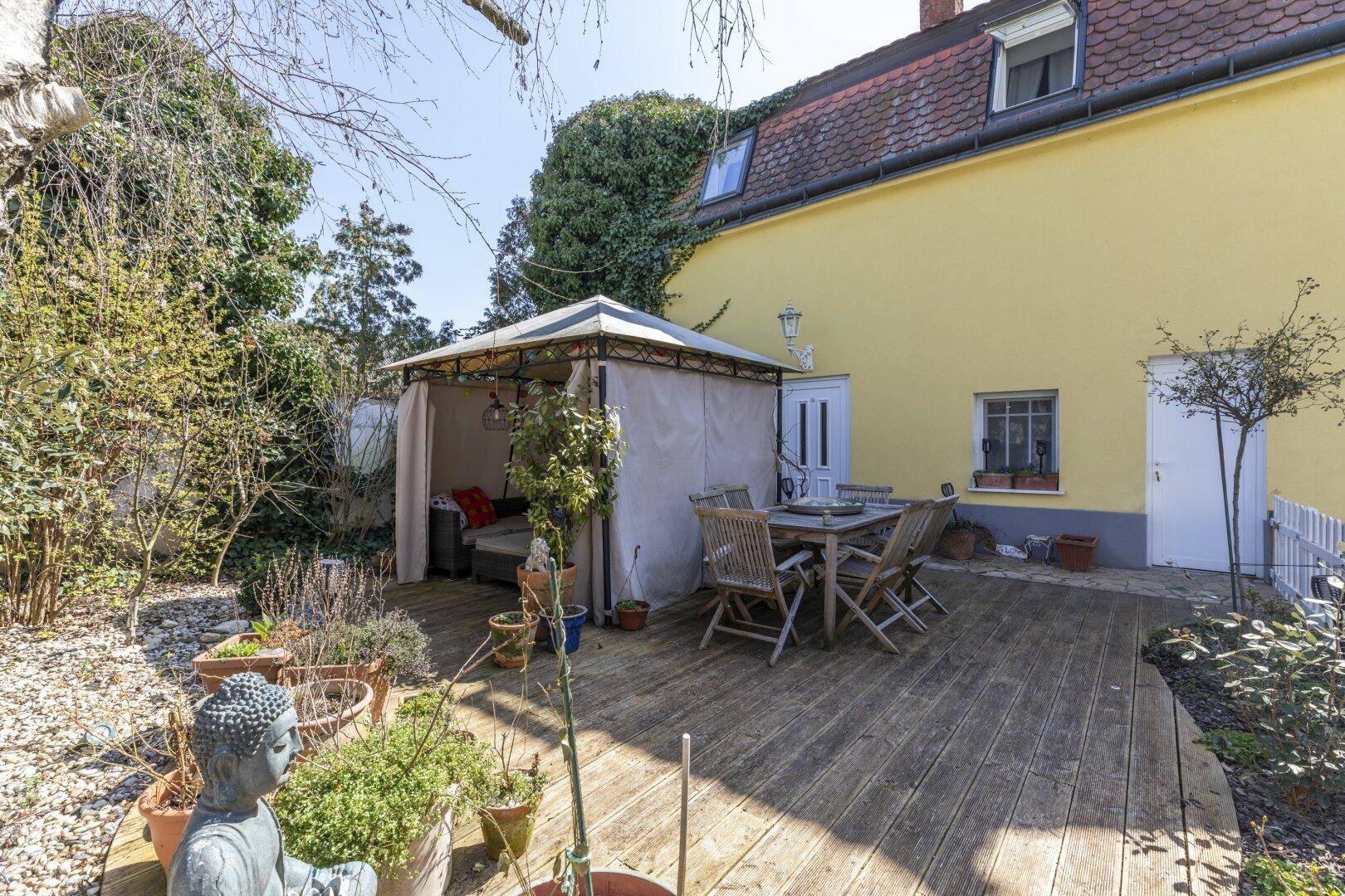 Holzterrasse mit Gartenmöbeln, Pavillon und Buddha-Statue im Grünen.