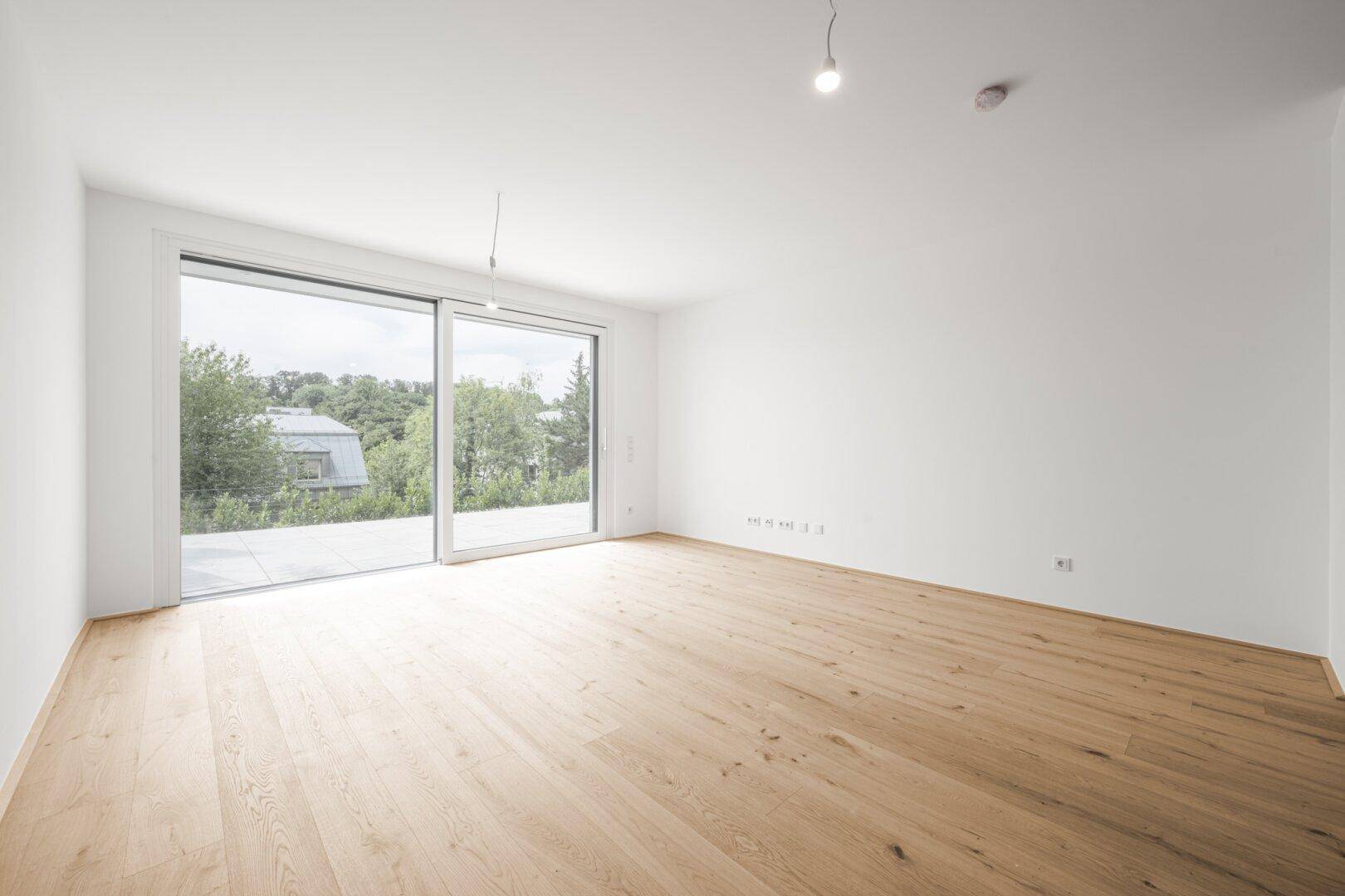 Heller, leerer Raum mit Holzboden und großer Fensterfront, die viel Licht hereinlässt.
