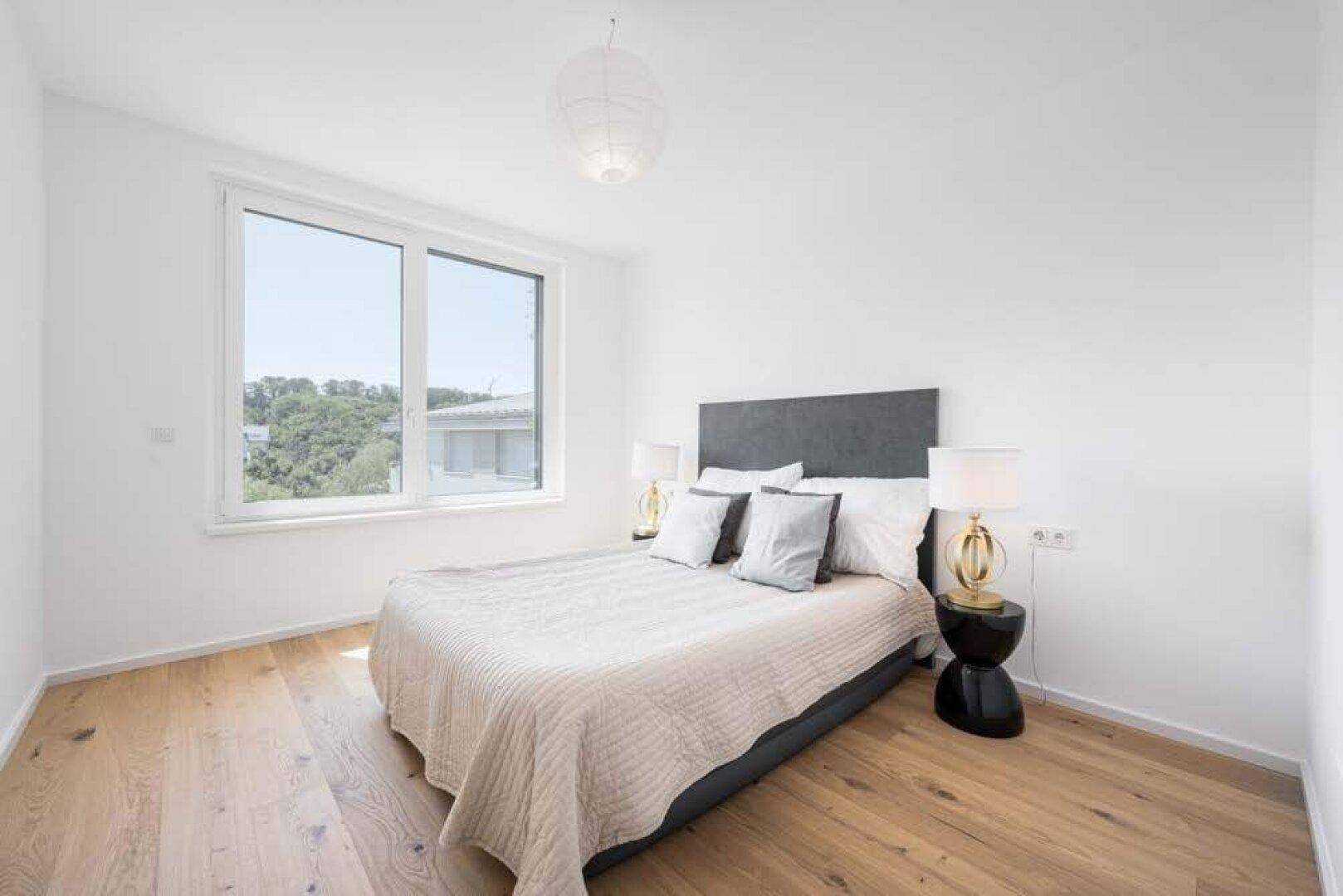 Helles Schlafzimmer mit Holzboden, Doppelbett und großem Fenster mit Grünblick.