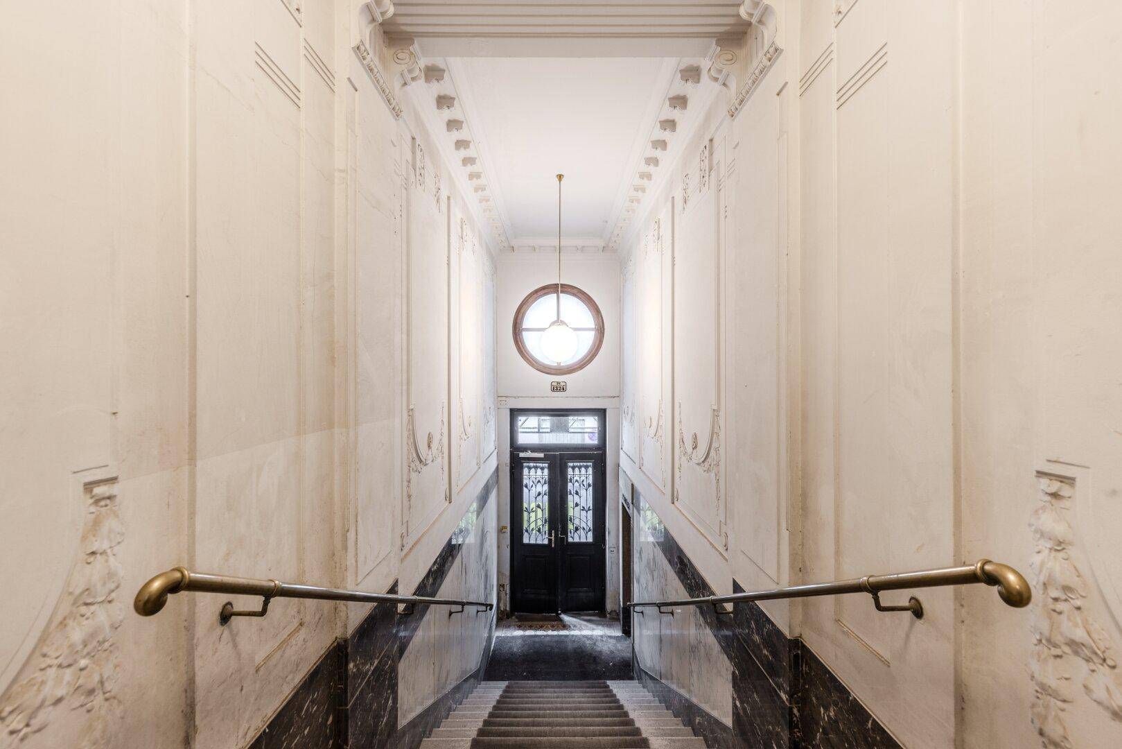 Elegantes Treppenhaus mit Marmorverkleidung, Stuckverzierungen und einem runden Fenster über dem Eingang.