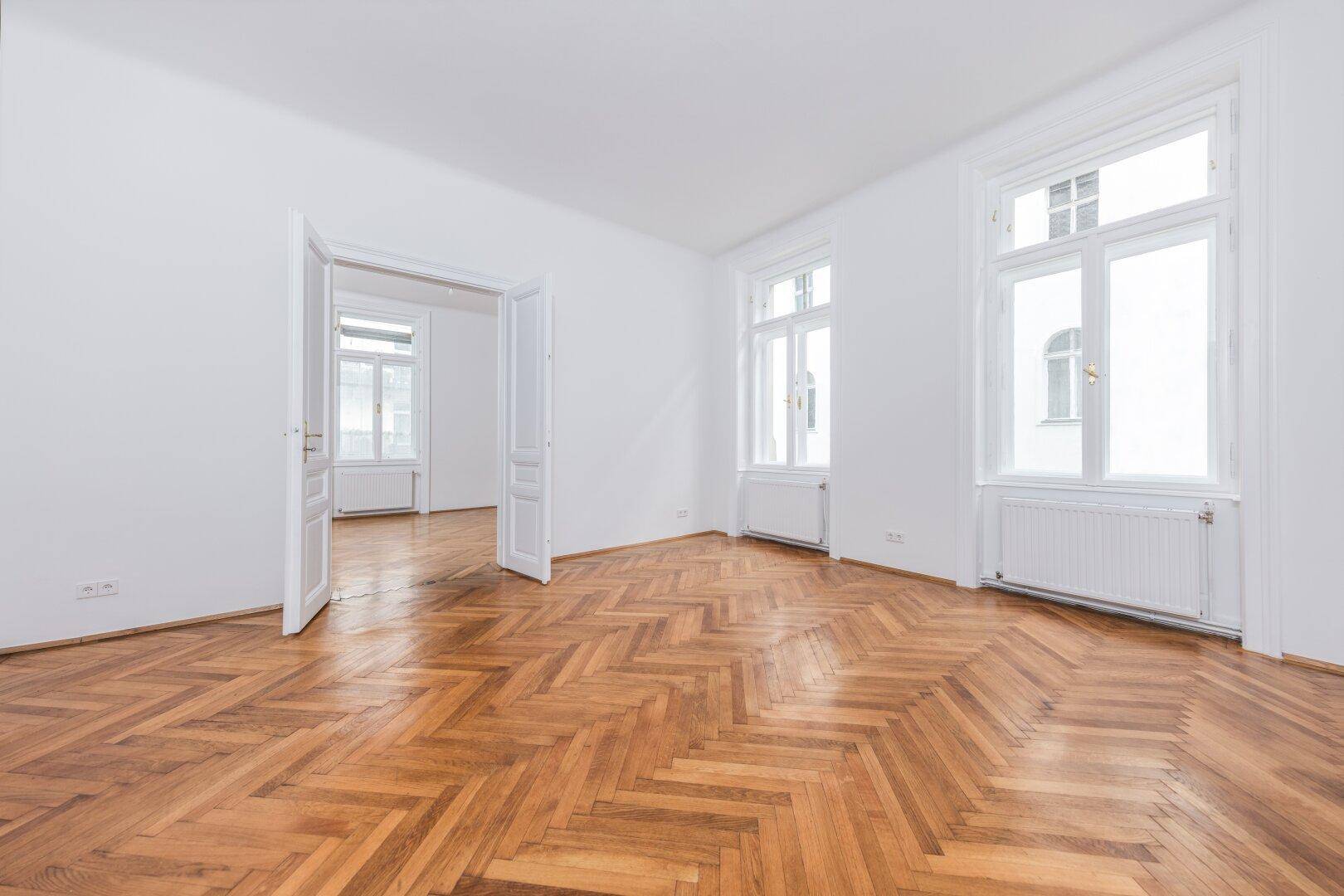 Geräumiges Zimmer mit Fischgrätparkett, hohen Decken und zwei großen Fenstern für viel Licht.