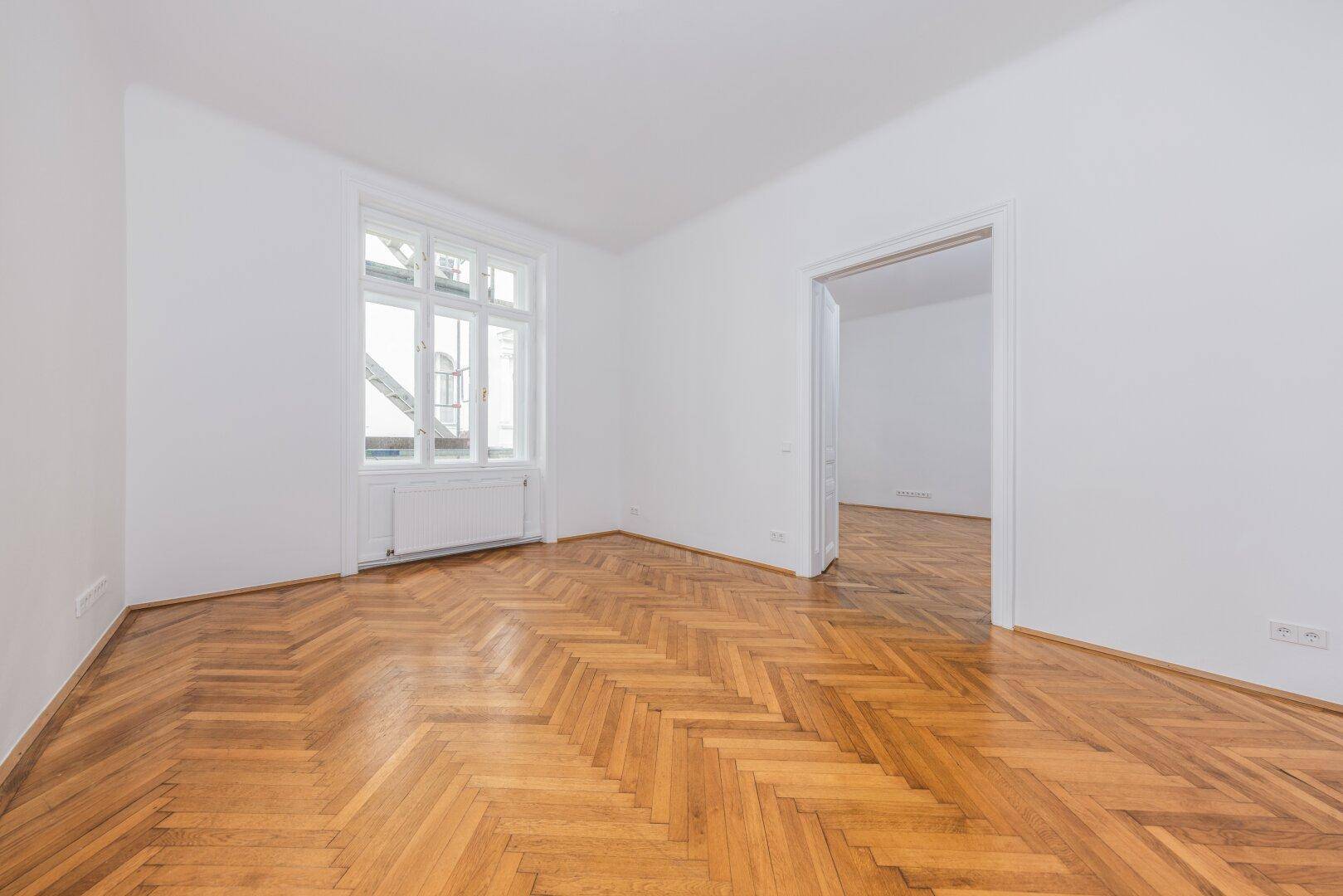 Helles Zimmer mit Fischgrätparkett, weißen Wänden und einem großen Fenster, das viel Tageslicht hereinlässt.