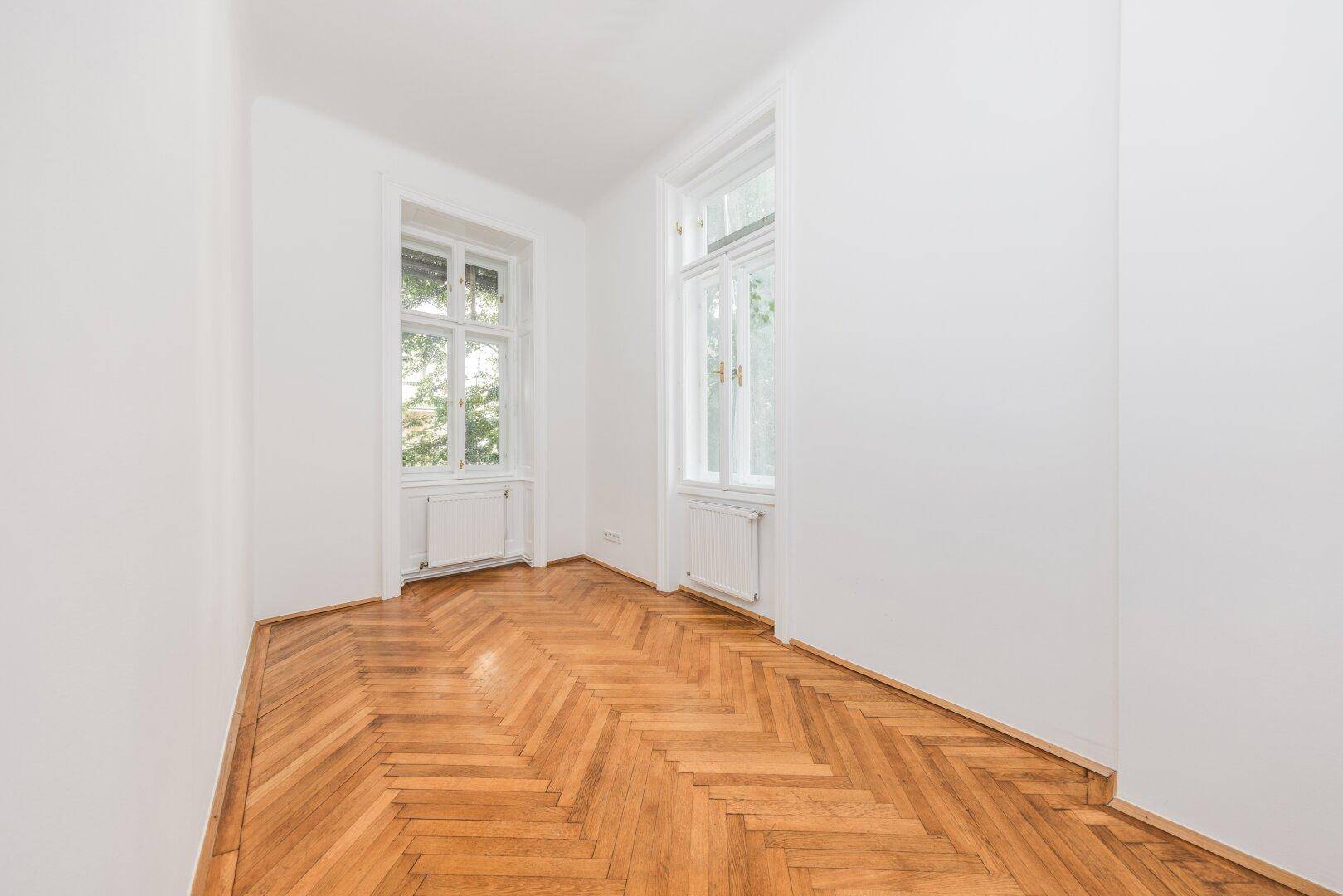 Ruhiges Zimmer mit Fischgrätparkett, weißen Wänden und zwei Fenstern, die eine grüne Aussicht bieten.