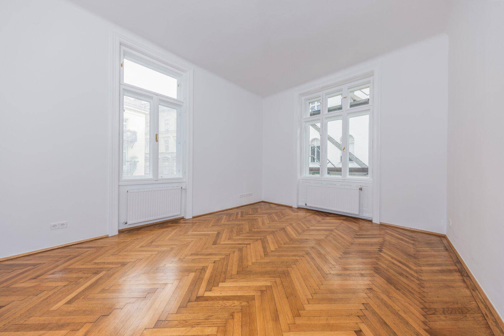 Großzügiges Zimmer mit Fischgrätparkett, weißen Wänden und zwei hohen Fenstern.