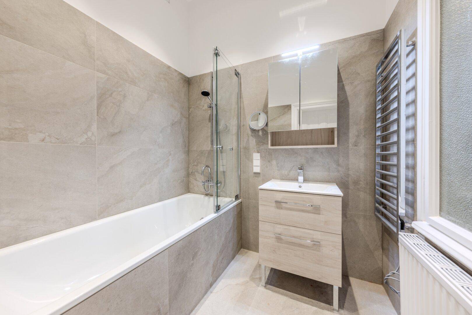 Modernes Badezimmer mit hellgrauen Fliesen, Badewanne mit Duschwand und beheiztem Handtuchhalter.