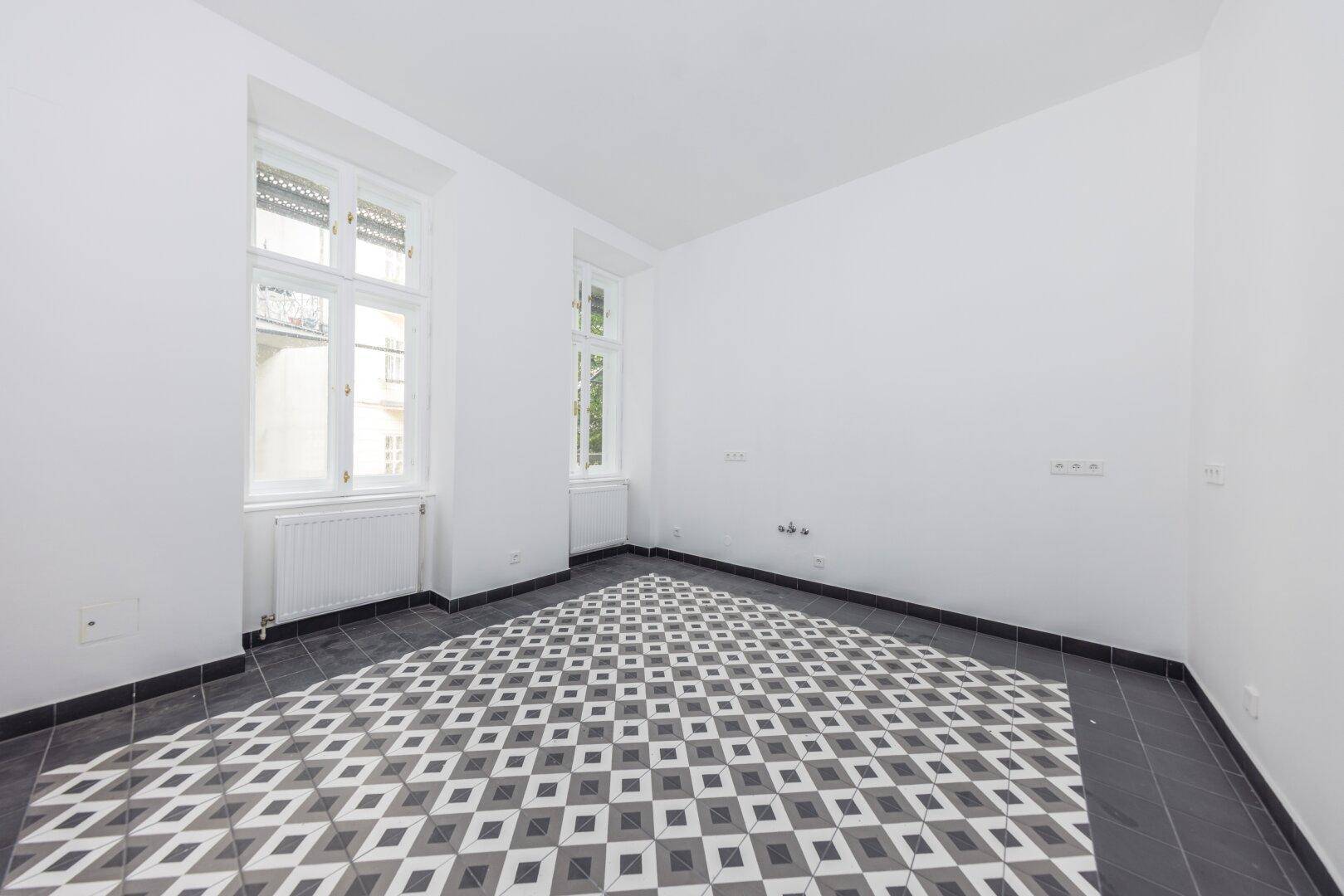 Heller Raum mit geometrischen Bodenfliesen, weißen Wänden und zwei Fenstern, ideal für eine Küche.