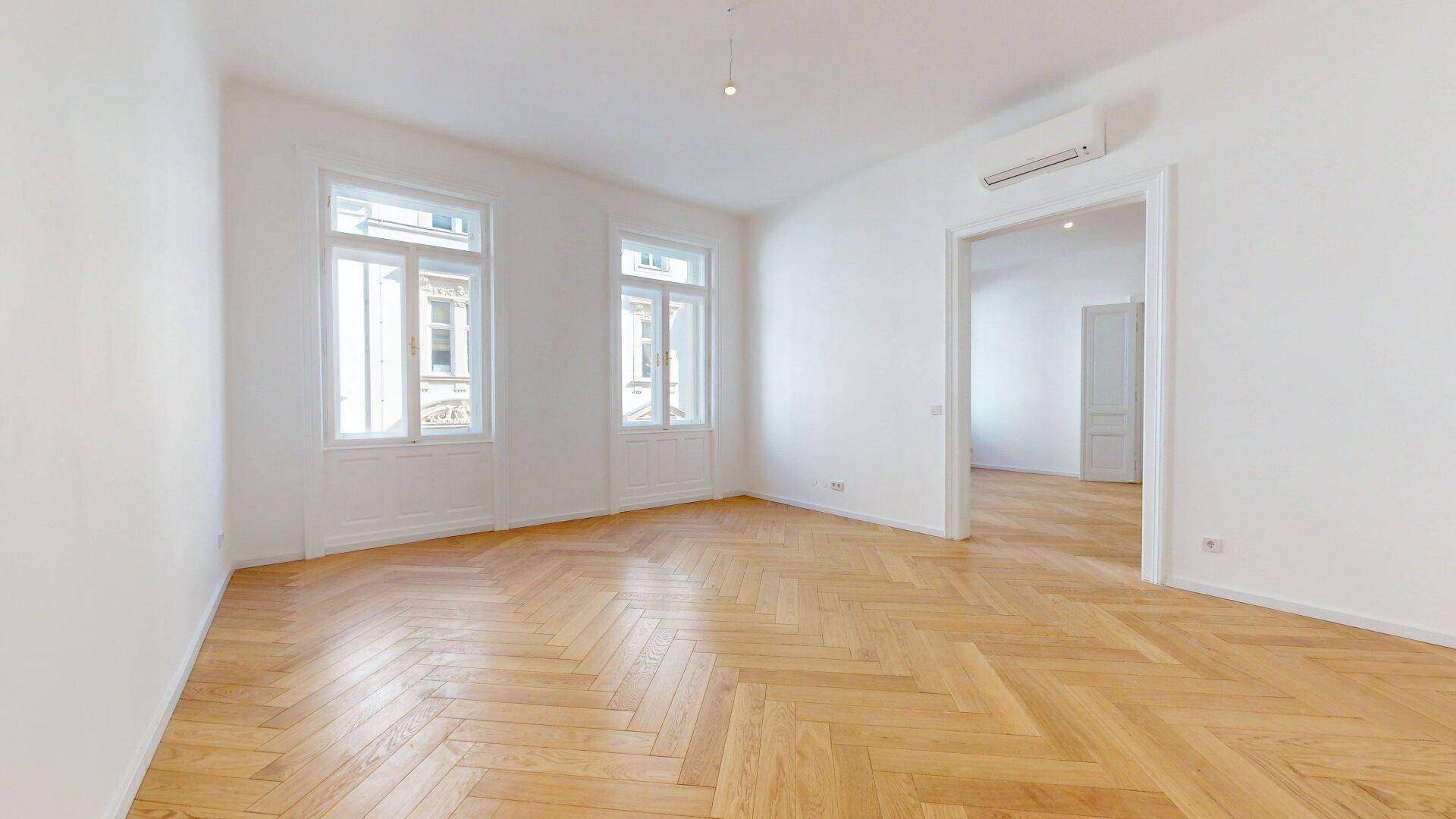 Helles und geräumiges Zimmer mit Fischgrätparkett, Klimaanlage und zwei hohen Fenstern.