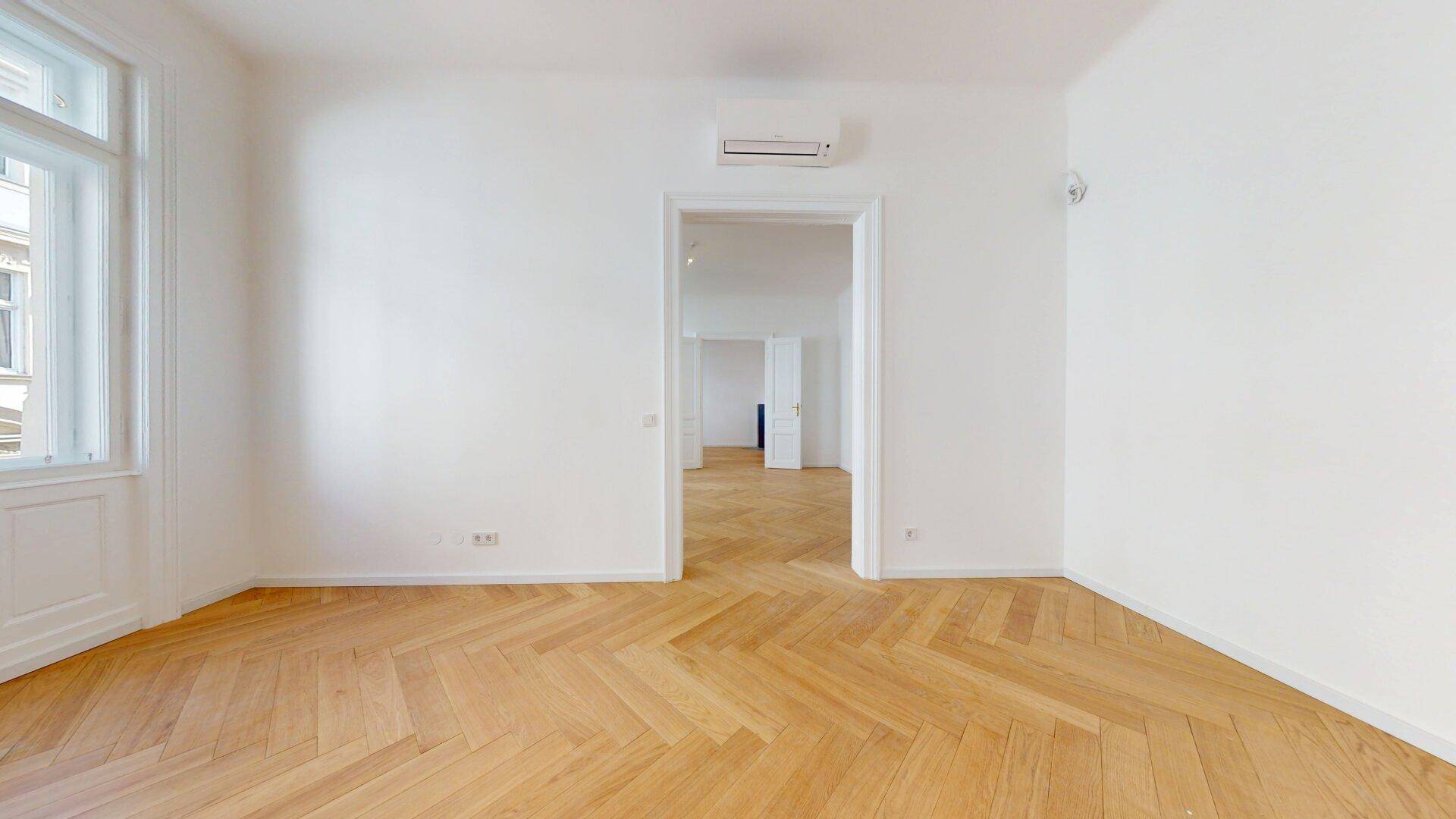 Geräumiges Zimmer mit Fischgrätparkett, Klimaanlage und einem großen Fenster, das viel Tageslicht bietet.