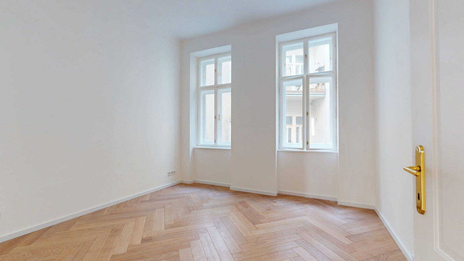Helles Zimmer mit Fischgrätparkett und zwei hohen Fenstern, das eine angenehme Wohnatmosphäre schafft.